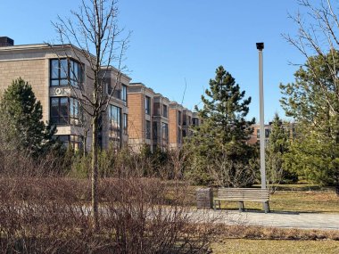 Modern, çok katlı, büyük pencereli ve bej taşlı cepheli binalar. Mimari zarif ve çağdaş. Hava açık ve güneşli, açık mavi bir gökyüzü var.