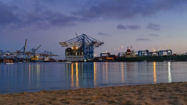 Geceleyin hareketli liman sahnesinde kargo gemileri, aydınlanmış vinçler ve kumlu kıyı şeridinden bakıldığında sakin su üzerindeki yansımaları görülüyor.