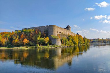 İvangorod Kalesi 'nin renkli sonbahar ağaçlarıyla çevrili manzarası açık mavi gökyüzünün altındaki sakin nehirde yansıyor.