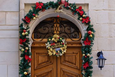 Noel çelenkleri, çelenkler, süsler ve taş bina cephesindeki şenlikli ışıklarla süslenmiş ahşap kapı.