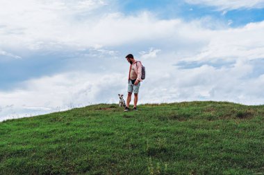 Sırt çantalı bir adam, bulutlu mavi gökyüzünün altındaki yeşil tepede küçük bir köpekle duruyor.