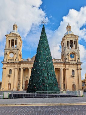 Christmas tree stands in front of historic Maltese church with twin bell towers, decorated for festive season under bright blue sky clipart