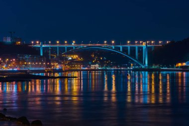 Gece aydınlık köprü, sakin nehre renkli yansımalar yapıyor. Parlayan şehir ışıkları ve karanlık şehir silüeti ile çevrili.