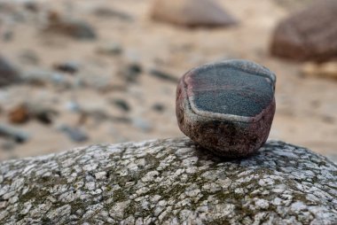 Vibrant multicolored stone resting on weathered rock surface, set against blurred sandy beach backdrop for natural coastal vibe clipart