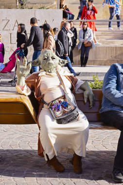 Street performer dressed as Yoda relaxing on bench, giving a hand five gesture, creating a humorous and surreal urban scene: Valletta, Malta - February 23, 2020 clipart