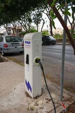 Şehir kaldırımında park halindeki arabalar ve gür ağaçlar arasında yer alan aktif şarj kablosu olan modern elektrikli araç şarj istasyonu.