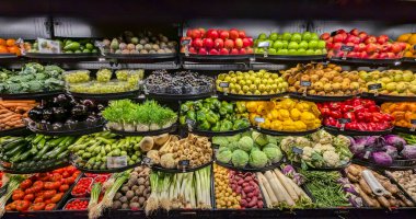 Renk ve tipine göre sıralanmış taze ürünlerin tam sergilendiği sergi biber, üzüm, elma, lahana ve daha fazlasını içeriyor. Süpermarketteki karanlık raflarla çerçevelenmiş görüntü