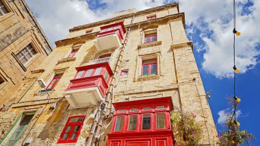 Mavi gökyüzünün altında sıcak kireçtaşı duvarlar üzerine istiflenmiş kırmızı ahşap balkonları olan güneşli Valletta köşesi.