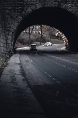 Bir kişi gece, asfalt yol yüzeyine gölgeler düşüren flaşlı fotoğrafçılıkla bir köprünün altında geziniyor. Tek renkli manzara, karanlık gökyüzüne karşı, su yansımaları ile kurulmuş.