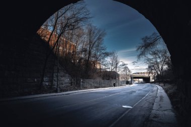 Bir kişi gece, asfalt yol yüzeyine gölgeler düşüren flaşlı fotoğrafçılıkla bir köprünün altında geziniyor. Tek renkli manzara, karanlık gökyüzüne karşı, su yansımaları ile kurulmuş.