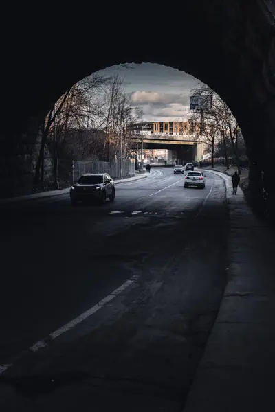 Bir kişi gece, asfalt yol yüzeyine gölgeler düşüren flaşlı fotoğrafçılıkla bir köprünün altında geziniyor. Tek renkli manzara, karanlık gökyüzüne karşı, su yansımaları ile kurulmuş.