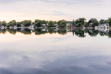 Arka planda ağaçlar olan bir gölde kayalık bir kıyı şeridi, bulutlu bir gökyüzünün altındaki suların kenarında doğal bir manzara tasvir ediyor.