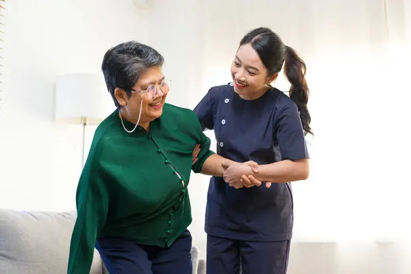 Female Asian nurse help old lady stand up from a couch.