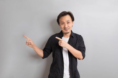 Siyah tişörtlü Asyalı bir adamın portresi gri stüdyo arka planına karşı duruyor ve solu işaret ediyor.