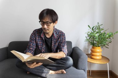 Asyalı adam kanepede oturmuş kitap okuyor.