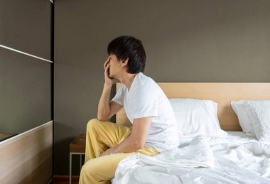 Beyaz tişörtlü üzgün Asyalı adam yatakta tek başına oturuyor ve bir şeyler düşünüyor..