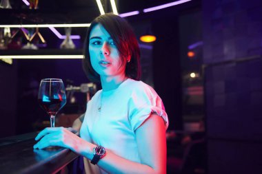 Kahverengi saçlı güzel bir kadın gece kulübünde dikiliyor ve elinde içki tutuyor..