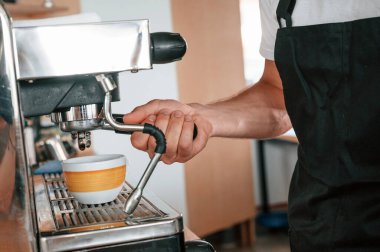 Kahve makinesini kullanarak. Beyaz gömlekli ve siyah önlüklü kafe çalışanı içeride..
