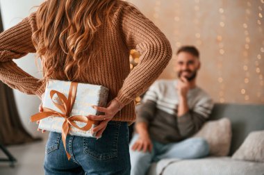 Kadın sürpriz yapıyor, elinde hediye kutusu var. Güzel genç çift yeni yılı evde kutluyor..