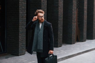 Siyah renkli giysi ve çanta. Sakallı ve gözlüklü şık adam binanın yanında..