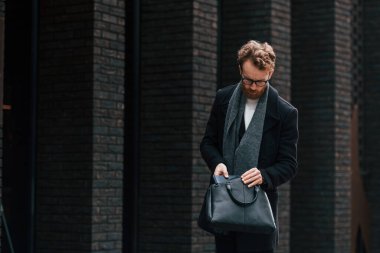 Siyah renkli giysi ve çanta. Sakallı ve gözlüklü şık adam binanın yanında..