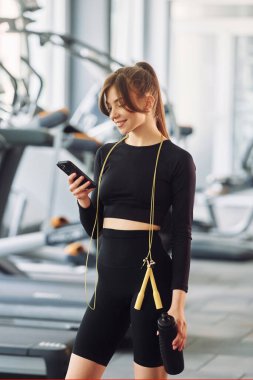 Ellerinde telefon ve boynunda ip ile. Spor elbiseli genç bir kadın spor salonunda spor yapıyor..