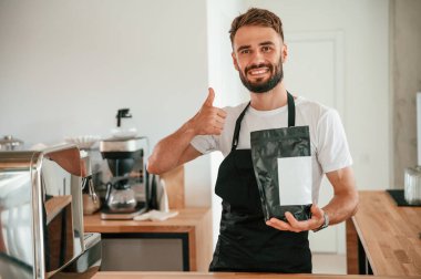 Elinde çiğ kahve paketi var ve parmağını kaldırıyor. Beyaz gömlekli ve siyah önlüklü kafe çalışanı içeride..