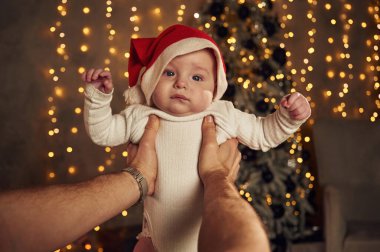 Adam elinde Noel Baba şapkalı küçük bir bebek tutuyor. İçerde, yeni yıl çelenkleri arkasında.