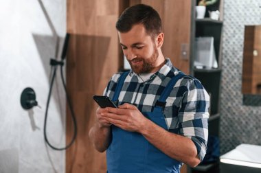 Ayakta ve akıllı telefonu tutarak. Mavi üniformalı tesisatçı banyoda çalışıyor..