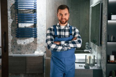 Kollarını kavuşturmuş duruyorsun. Mavi üniformalı tesisatçı banyoda çalışıyor..