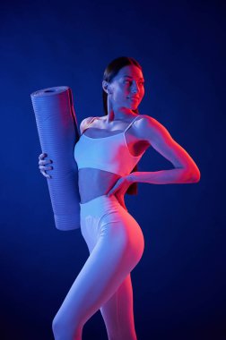 Egzersiz için hazır. Yoga minderiyle. Spor elbiseli genç bayan stüdyoda neon ışıklarıyla bekliyor..