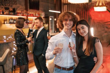 Kadın ve erkek ayakta duruyor ve poz veriyor. Güzel giyinmiş bir grup insan içeride yeni yılı kutluyorlar..