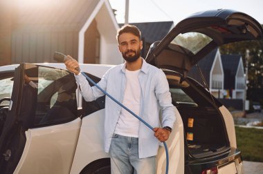 Güzel güneş ışığına karşı. Şarj kabloları duruyor. Genç şık adam, gündüz vakti elektrikli arabayla geziyor..