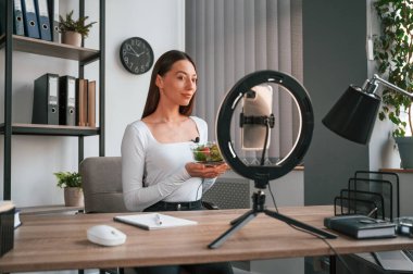 Beslenme uzmanı kamerada salata gösteriyor. Beyaz elbiseli genç ve güzel bayan ofiste..