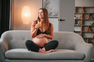 Elinde portakal suyuyla. Kanepede. Güzel hamile kadın evde..