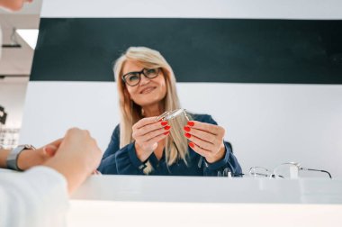 Masanın yanında oturuyor. Mağazadan gözlük seçen ve asistanından yardım alan bir kadın.