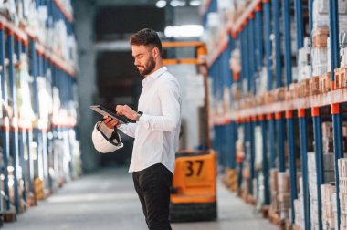 Ayakta durup tableti tutarak. Depo işçisi depoda bir sürü ürünle birlikte..