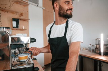 Kahve makinesini kullanarak. Beyaz gömlekli ve siyah önlüklü kafe çalışanı içeride..