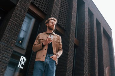 Telefonla. Sakallı, haki renkli ceketli ve kot pantolonlu şık bir adam binanın yanında..