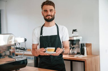 Kupa ellerde. Beyaz gömlekli ve siyah önlüklü kafe çalışanı içeride..