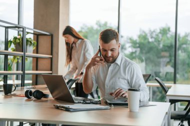 Patron şu an telefonda konuşuyor. Erkek ve kadın modern ofiste birlikte çalışıyorlar..