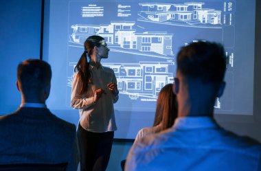 İşçilerle konuşan kadın lider, borsa şirketinin projektörüyle ilgili planı gösteriyor..