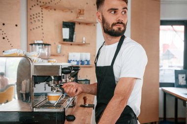 Kahve makinesini kullanarak. Beyaz gömlekli ve siyah önlüklü kafe çalışanı içeride..