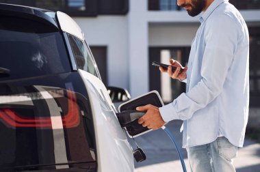 Aracı şarj etmek ve telefon kullanmak. Genç şık adam, gündüz vakti elektrikli arabayla geziyor..