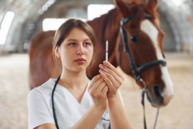Bir hıyara şırınga hazırlıyorum. Beyaz önlüklü kadın doktor ahırda atıyla birlikte..