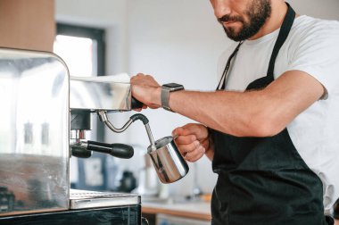 Ayakta ve kahve doldururken. Beyaz gömlekli ve siyah önlüklü kafe çalışanı içeride..