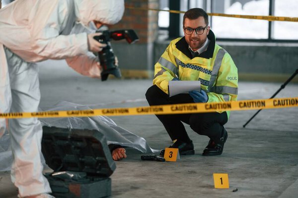 Женщина делает фотографии с помощью камеры. Детективы собирают улики на месте преступления рядом с трупом.
