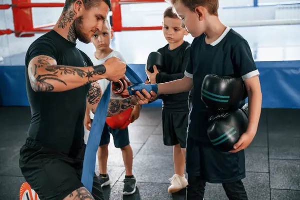 stock image Wearing blue hand wraps. Young tattooed coach teaching the kids boxing techniques.