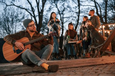 Gitarlı bir adam arkadaşlarının önünde oturuyor. Bir grup insan arka bahçede birlikte vakit geçiriyor..