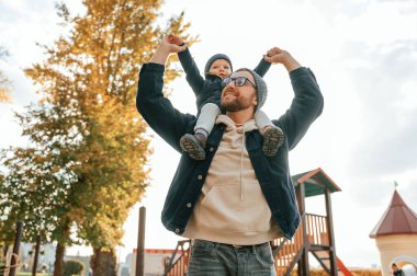 Çocuk insanın omuzlarında oturuyor. Baba ve küçük oğlu gündüz vakti dışarıda beraberler..